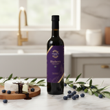 Tastefully Olive Blueberry dark balsamic bottle with blueberries, olive branches, and cheese drizzled with balsamic on a kitchen counter.