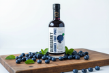 Tastefully Olive Blueberry crushed fruit puree bottle surrounded by blueberries and mint on a wooden board.