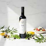 Tastefully Olive Greek Seasoning white balsamic bottle with lemon, olives, herbs, and a bowl of Greek salad.