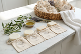 Tastefully Olive handmade soaps and drawstring pouches on marble with a woven basket, olive branches, lavender, and a bowl of oil.