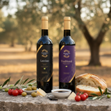 Tastefully Olive Leccino olive oil and Traditional dark balsamic bottles with bread, cheese, tomatoes, olives, and a dipping bowl in an olive grove.