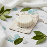 Round cream-colored soap on a Mykonos drawstring pouch with green leaves and blue sea glass on marble.