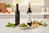 Tastefully Olive Pesto olive oil and Peach white balsamic bottles with pesto pasta, basil leaves, and peach slices in a bright kitchen.