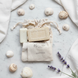 Rectangular cream-colored soaps with Greek-key borders on Tastefully Olive Santorini pouches surrounded by seashells and lavender.