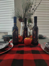 Three Tastefully Olive Red Apple dark balsamic bottles with red apples and a vase of branches on a checked table runner.