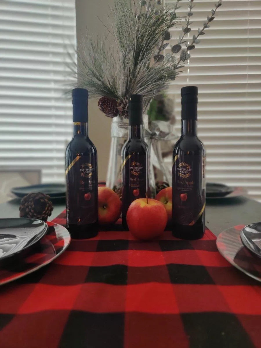 Three Tastefully Olive Red Apple dark balsamic bottles with red apples and a vase of branches on a checked table runner.