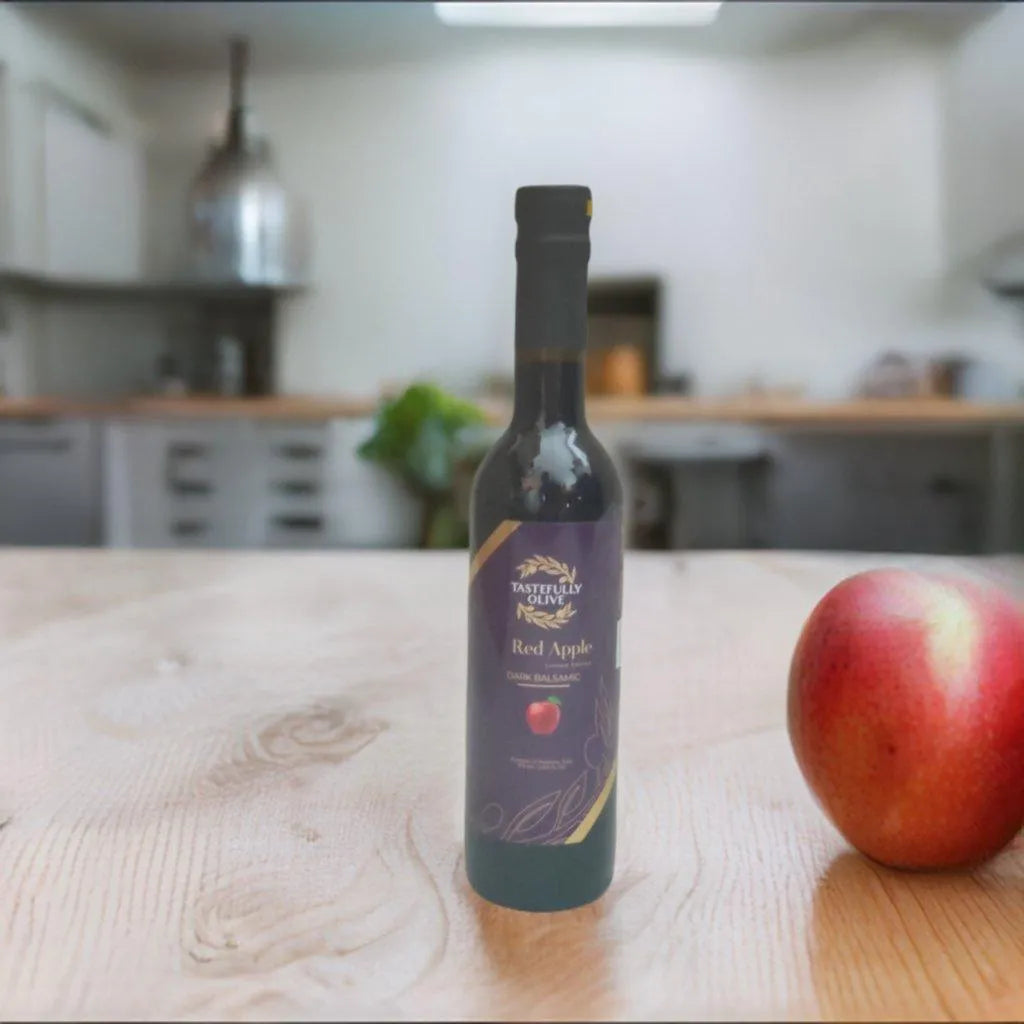 Tastefully Olive Red Apple dark balsamic bottle beside a red apple on a wooden kitchen counter.