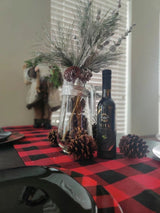 Tastefully Olive Vita olive oil bottle with pinecones and a vase of branches on a red-and-black checked table runner.